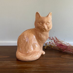 May include: A ceramic figurine of a light brown cat sitting with its head tilted slightly to the side. The cat has a detailed, textured fur pattern.