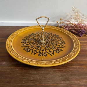 Puede incluir: Una bandeja de servir de dos niveles con asa dorada. La bandeja es de color amarillo mostaza con un diseño floral negro en el centro. El borde de la bandeja tiene un diseño festoneado. Una pieza decorativa para servir alimentos o exhibir artículos.