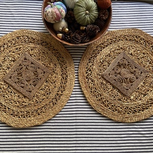 May include: Two round, woven, brown placemats with a decorative, intricate design in the centre. The placemats are on a striped, black and white surface.