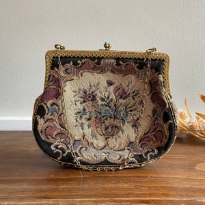 Puede incluir: Un bolso vintage de tapicería con un diseño floral y una cadena dorada. El bolso está cerrado y tiene una pequeña piedra turquesa en la parte superior.