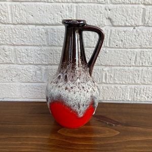 May include: A vintage ceramic pitcher with a handle. The pitcher features a dark brown top, a white and brown speckled middle, and a bright red base. The pitcher is set against a white brick wall and a wooden surface.
