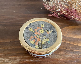 Könnte beinhalten: Eine runde, weiße Schmuckschatulle mit goldfarbenem Rand und einem floralen Design auf dem Deckel. Das Design zeigt orange, gelbe und grüne Blumen mit einem Vogel im Flug.