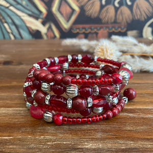 May include: A spiral wrap bracelet with red beads and silver-colored accents. The bracelet is made of various sizes and shapes of beads, creating a layered effect. The beads are a deep red color, and the bracelet is wrapped around itself.