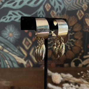 Könnte beinhalten: Goldfarbene Creolen mit hängenden, strukturierten, tropfenförmigen Anhängern. Die Ohrringe werden auf einem schwarzen Ständer vor einem Hintergrund aus gemusterter Tapete präsentiert. Ein stilvolles Accessoire.