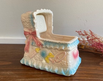 May include: A white ceramic baby crib planter with blue and pink accents. The crib has a pink bow, blue trim, and a heart-shaped design on the side. The planter is decorated with baby-themed details, including a rattle, a pacifier, and a baby shoe.