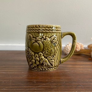 May include: A green ceramic mug with a raised design of fruit and leaves. The mug has a handle and a rope-like design around the top.