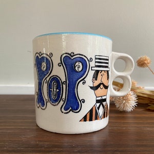 Puede incluir: Taza de cerámica blanca con un diseño azul y negro. La taza tiene la palabra "POP" escrita en una fuente estilizada y presenta un hombre de dibujos animados con bigote y sombrero de copa. La taza tiene dos asas.
