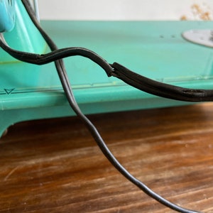 May include: A vintage turquoise sewing machine with a black electrical cord. The cord is frayed and worn.