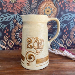 以下が含まれることがあります： ベージュ色のセラミックピッチャー。ハンドル付きで、「2015」の数字と茶色の水平ストライプのエンボス加工が施されています。木製の表面に置かれています。