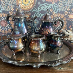 May include: A vintage silver tea set, comprising a teapot, creamer, sugar bowl, and serving tray. The set has intricate detailing and a tarnished finish, with iridescent colours. The tray is oval-shaped with a decorative edge. The background features patterned wallpaper.
