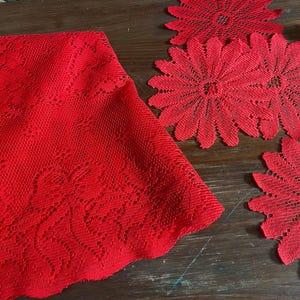 May include: A red lace tablecloth with a floral pattern is displayed alongside four matching red daisy-shaped doilies. The items are arranged on a dark wooden surface, creating a contrast of textures and colors.