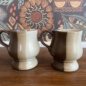 Puede incluir: Dos tazas de cerámica con un diseño único. Cada taza está dividida verticalmente en dos secciones de bloques de color: un lado es de color beige claro y el otro es marrón oscuro. Las tazas tienen una base redondeada y un asa curva.