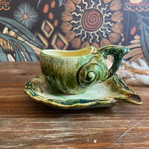 May include: A ceramic teacup and saucer set with a seashell design. The cup and saucer are glazed in shades of green and yellow. The teacup has a handle and sits on a matching saucer. The set is on a wooden surface.