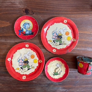 May include: A set of four red and white toy plates and a red and blue toy cup with a bunny and cat design. The plates and cup are made of metal and have a vintage look.