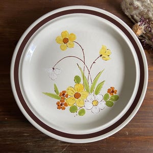 May include: Round, off-white ceramic plate with a brown rim and floral design. The flowers are yellow, white, and orange with green leaves. A brown band encircles the edge of the plate.