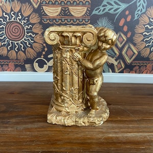 May include: Gold-toned decorative column with a cherub sculpture. The cherub is holding onto the column and has a vine-like design on the base.