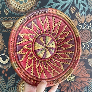 Può includere: Un cesto rotondo intrecciato con un vivace design geometrico. Il cesto presenta un motivo circolare centrale nei toni del viola, verde e giallo, circondato da segmenti rossi e dorati radianti. Il bordo è rosso scuro.