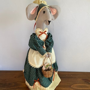 May include: A gray mouse doll wearing a green dress with white lace trim and a red bow. The doll is holding a small wicker basket filled with pink flowers.