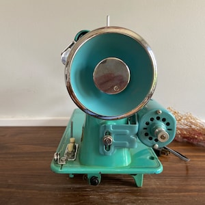 May include: A vintage turquoise blue sewing machine with a chrome-plated bobbin winder and a metal foot pedal. The machine has a white plastic knob and a metal lever.