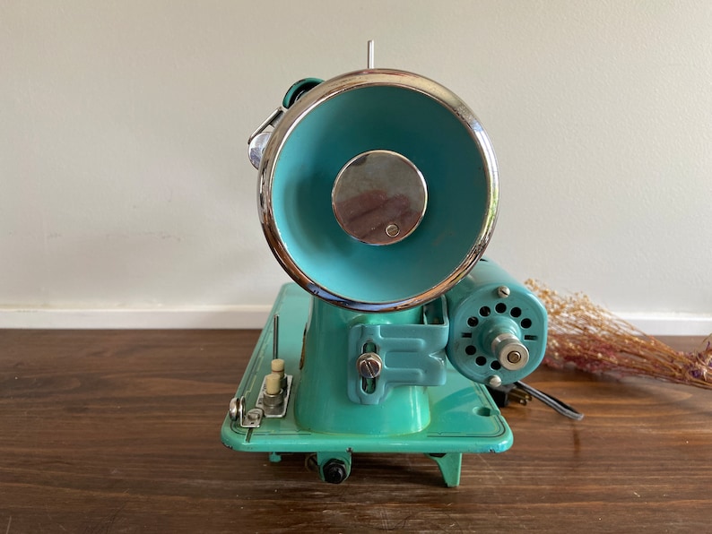 May include: A vintage turquoise blue sewing machine with a chrome-plated bobbin winder and a metal foot pedal. The machine has a white plastic knob and a metal lever.