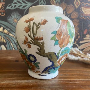 May include: Cream-colored ceramic vase with floral designs in shades of orange, green, and blue. The vase features a round body and a short, flared neck. The floral pattern includes branches and flowers.
