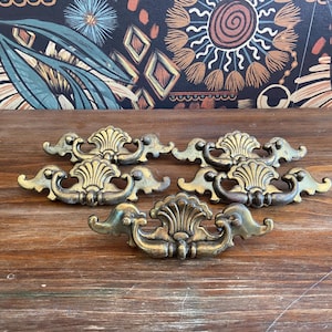 May include: Five antique brass drawer pulls with a shell and scroll design. The pulls are arranged on a wooden surface, showcasing their aged patina. The background features a patterned fabric with floral and geometric elements.