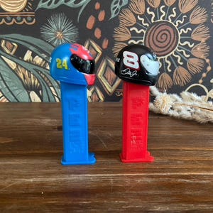以下が含まれることがあります： レーシングヘルメットのデザインが施された2つのPEZディスペンサー。1つは24番の青色、もう1つは8番とサインの赤色です。どちらも黒いバイザーで、木製の表面に置かれています。