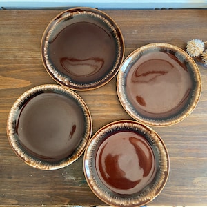 Pode incluir: Quatro pratos de cerâmica castanhos com acabamento esmaltado. Os pratos têm um centro castanho escuro e uma borda castanho claro.