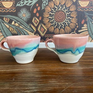 Puede incluir: Dos tazas de cerámica con un esmalte rosa, blanco y azul. Las tazas tienen una forma redondeada y un asa en un lado.