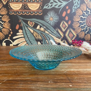 Puede incluir: Un cuenco de vidrio transparente de color turquesa con un patrón texturizado y punteado. El cuenco tiene una forma poco profunda y redondeada y se encuentra sobre una superficie de madera. Elementos decorativos en el fondo.