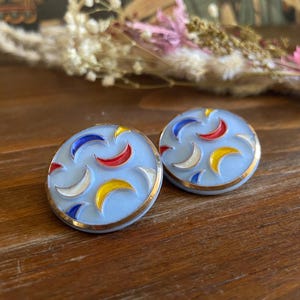 Puede incluir: Un par de pendientes redondos de color azul claro con un borde dorado. Cada pendiente presenta un patrón de formas de media luna coloridas en rojo, amarillo, blanco y azul. Los pendientes se exhiben sobre una superficie de madera.