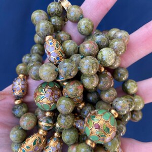 Puede incluir: Un collar de cuentas verdes y doradas con intrincados dise&ntilde;os florales. Las cuentas est&aacute;n hechas de una piedra verde moteada y se intercalan con cuentas doradas y cuentas redondas m&aacute;s grandes con coloridos patrones florales de esmalte.
