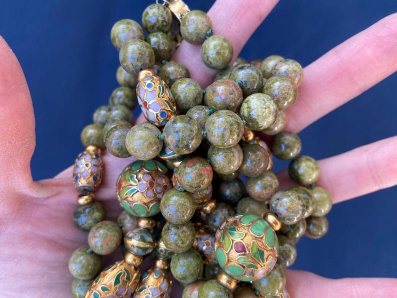 Puede incluir: Un collar de cuentas verdes y doradas con intrincados dise&ntilde;os florales. Las cuentas est&aacute;n hechas de una piedra verde moteada y se intercalan con cuentas doradas y cuentas redondas m&aacute;s grandes con coloridos patrones florales de esmalte.