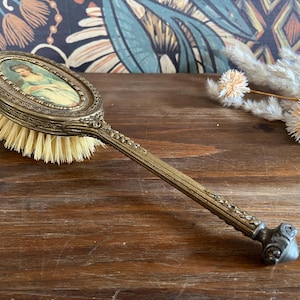 May include: A vintage gold-toned hairbrush with a decorative handle and a round mirror with a painted image of a woman in a dress. The brush has natural bristles.