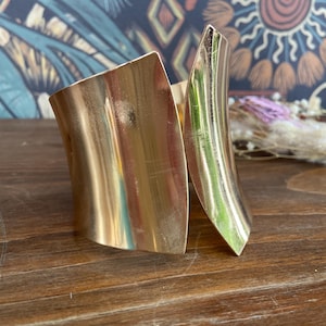 May include: A gold-colored cuff bracelet with a curved design. The bracelet has a wide, rectangular shape that wraps around the wrist, with a split design on one side. The metal has a polished finish, reflecting light.