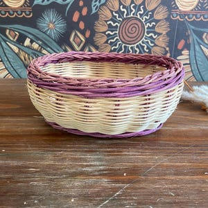 May include: A round woven basket with a cream-colored body and a purple rim. The basket is made of natural materials and sits on a wooden surface. The background features a patterned wallpaper.