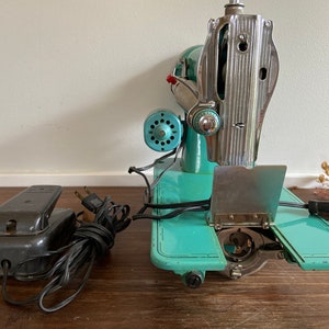 May include: A vintage turquoise and silver sewing machine with a foot pedal and cord. The machine has a metal plate with a hinged cover and a bobbin winder.