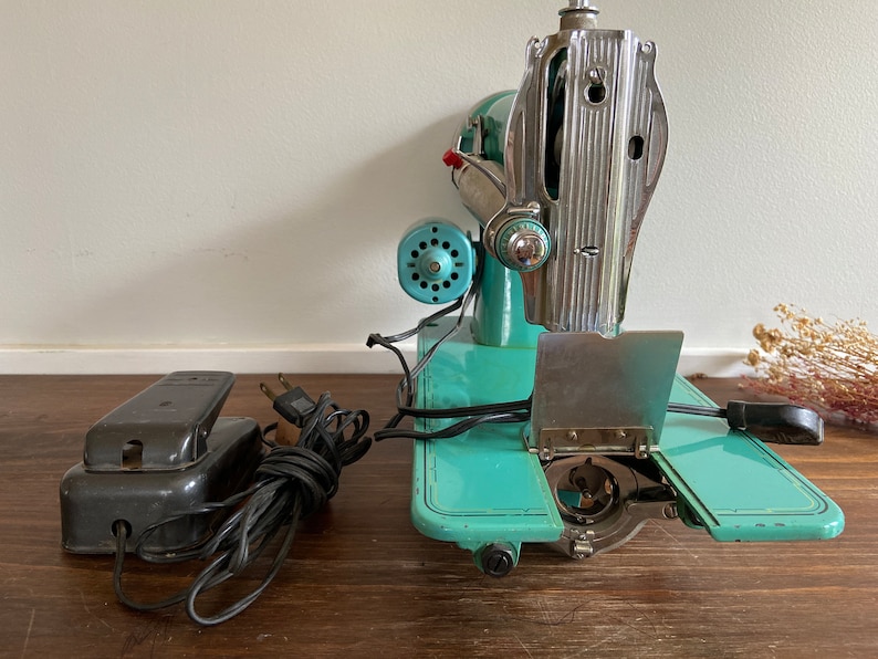May include: A vintage turquoise and silver sewing machine with a foot pedal and cord. The machine has a metal plate with a hinged cover and a bobbin winder.