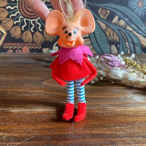 Puede incluir: Adorno de ratón vintage con cabeza color melocotón, cuello rosa y atuendo rojo. El ratón tiene piernas a rayas azules y blancas y botas rojas. El adorno es probablemente para un árbol de Navidad u otra exhibición festiva.