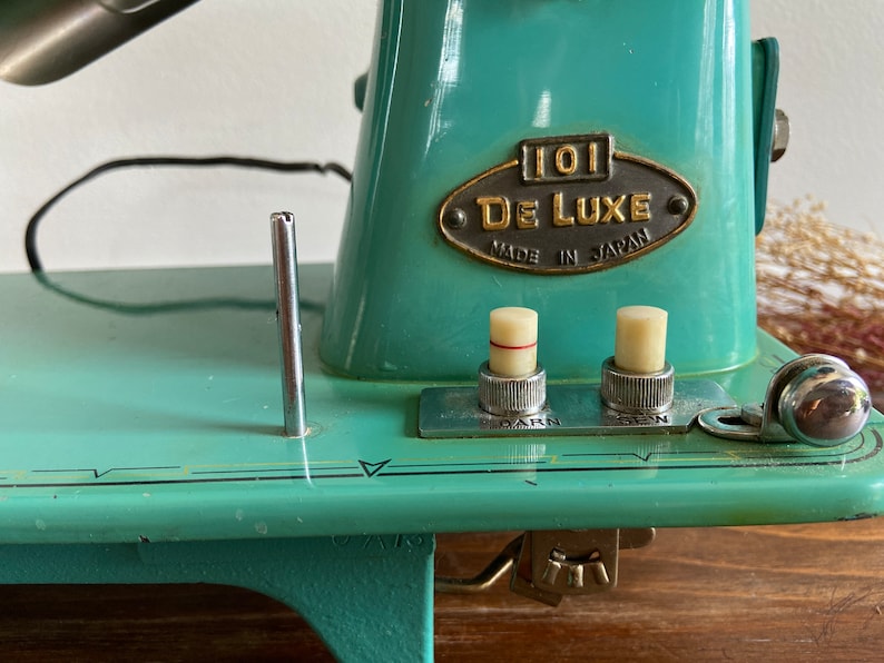 May include: A turquoise blue vintage sewing machine with a metal plate that reads "101 De Luxe Made in Japan". The machine has two knobs, one labeled "Darn" and the other labeled "Sew".