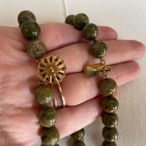 Puede incluir: Un collar de cuentas de piedra verde y marr&oacute;n con un cierre dorado y un colgante dorado en forma de flor.
