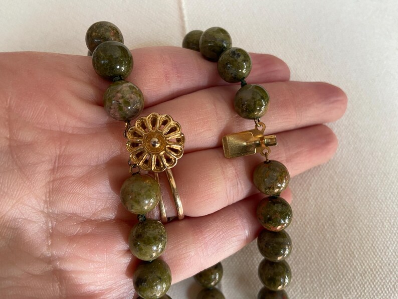 Puede incluir: Un collar de cuentas de piedra verde y marr&oacute;n con un cierre dorado y un colgante dorado en forma de flor.