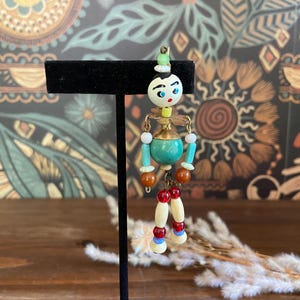 May include: A vintage articulated doll pendant with a painted wooden head, turquoise body, and dangling legs. The doll has a green hat, blue eyes, and red and white accents. It is suspended from a gold-toned metal chain.
