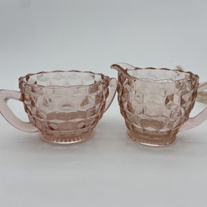 May include: A pair of vintage pink glass serving dishes. One is a sugar bowl with a handle, and the other is a creamer with a spout and handle. Both have a diamond pattern and are on a white background.