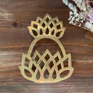 May include: A vintage brass trivet with a decorative openwork design. The trivet features a pineapple-like shape with a series of diamond and teardrop cutouts. The brass has a warm, aged patina. Dried flowers are in the background.