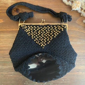May include: Black vintage handbag with a gold clasp and handle. The bag is crocheted with a decorative pattern of gold beads on the front. The bottom of the bag has a black, scalloped, decorative element.