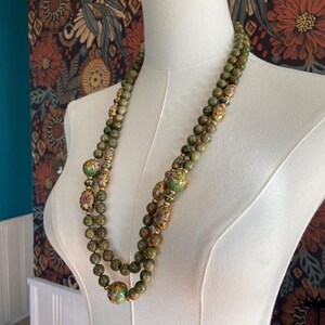 Puede incluir: Un collar de doble cadena hecho de cuentas de jade verde con detalles dorados y florales. El collar se muestra en un maniqu&iacute; blanco.