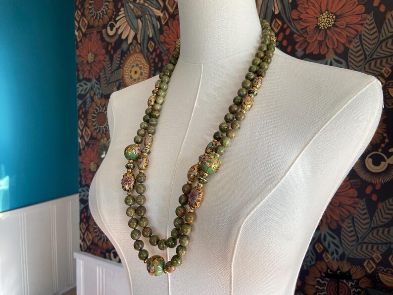 Puede incluir: Un collar de doble cadena hecho de cuentas de jade verde con detalles dorados y florales. El collar se muestra en un maniqu&iacute; blanco.
