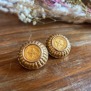May include: A pair of gold-toned Givenchy clip-on earrings. Each earring features a round, ridged design with a central medallion. The medallions display a starburst pattern and the Givenchy name. The earrings are set on a wooden surface.