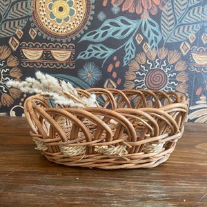May include: A woven wicker basket with a rounded oval shape. The basket is made of light brown wicker and has a decorative woven pattern. Dried grasses are placed inside the basket. The background features a dark floral wallpaper.